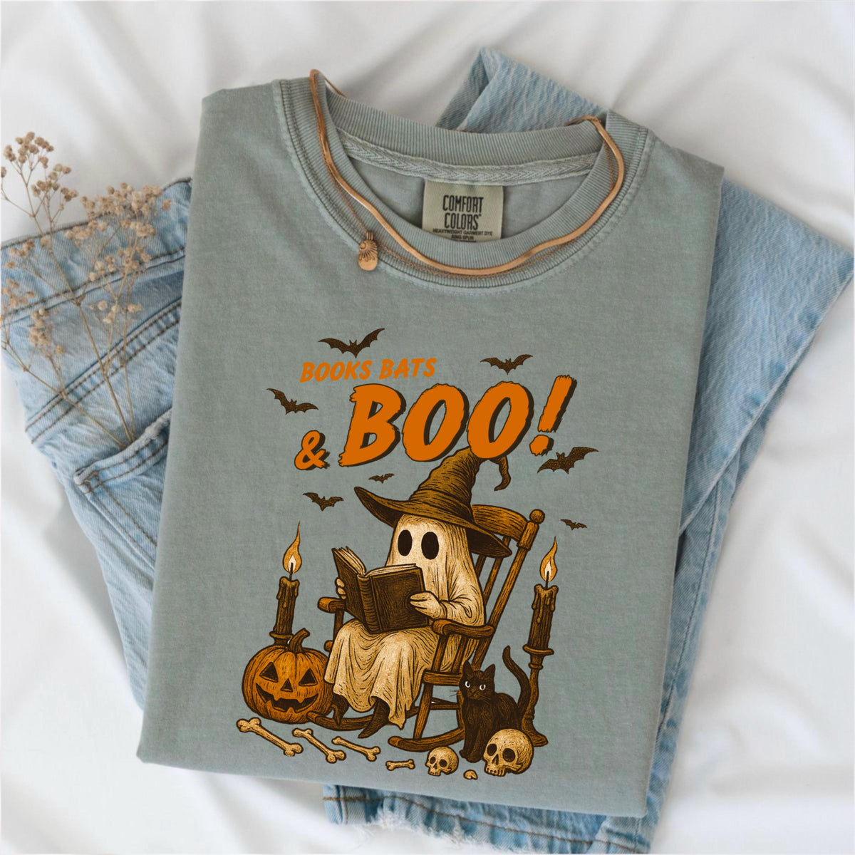 May include: Sage green Comfort Colors t-shirt with a Halloween-themed graphic. The design features a ghost reading a book in a rocking chair, with the text "Books Bats & Boo!" in orange. Includes bats, a pumpkin, candles, a black cat, bones, and skulls.