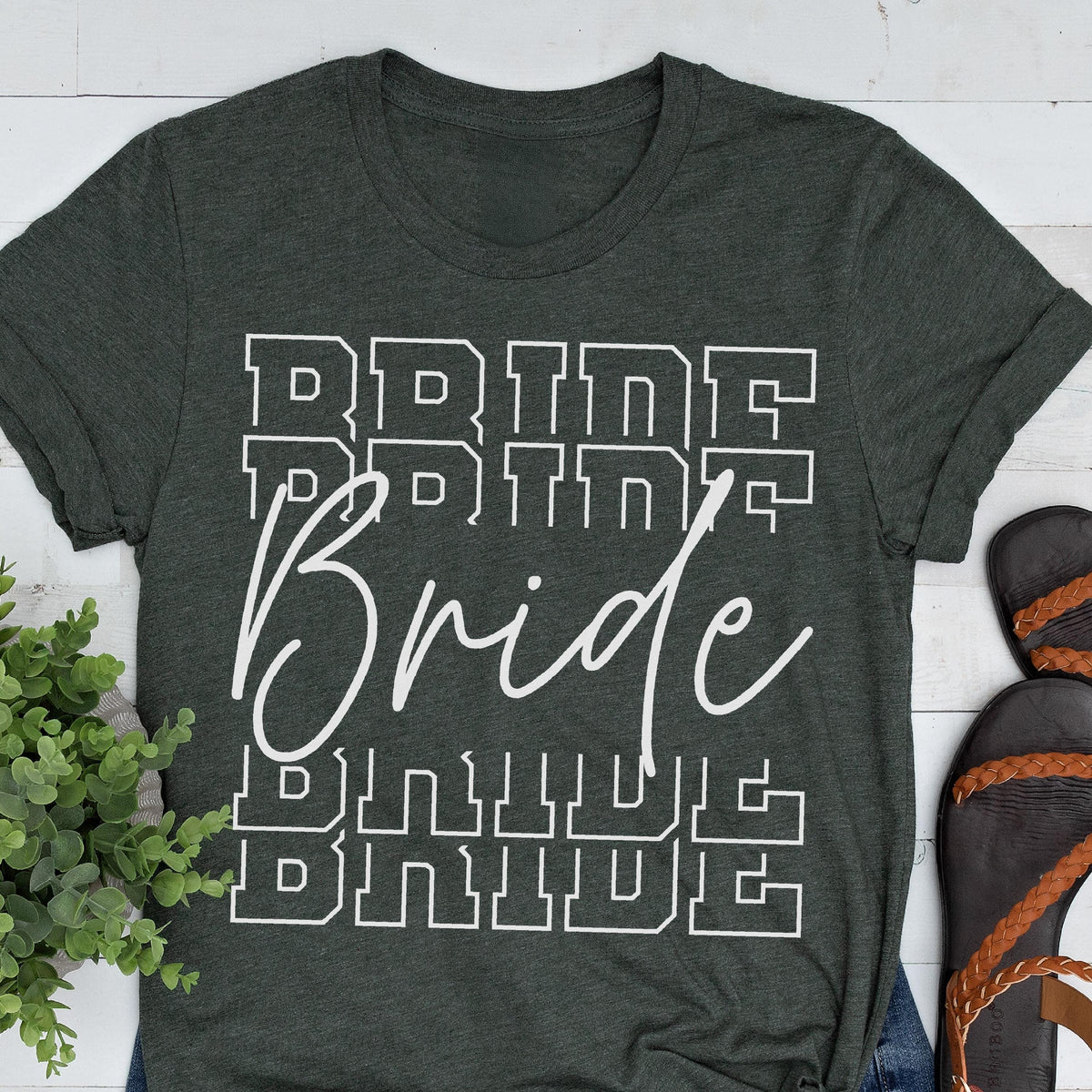 May include: Dark green t-shirt with the word "Bride" in a white script font, overlaid on the word "Bride" repeated in a block letter style. The shirt is folded at the sleeves.