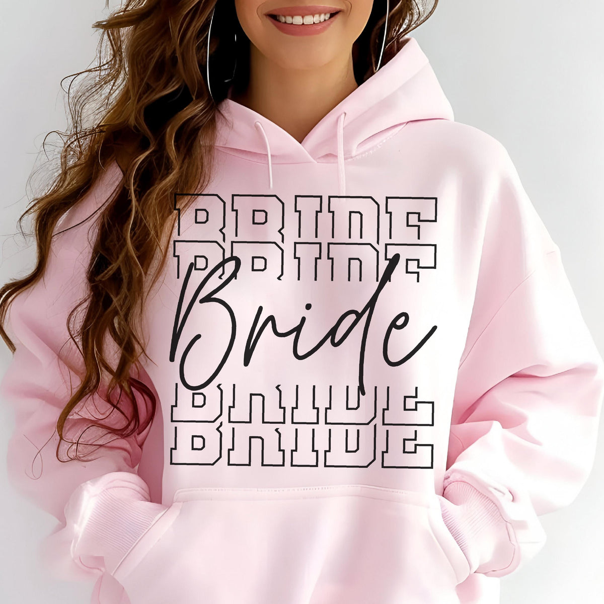 May include: A light pink hooded sweatshirt featuring a front graphic with the word Bride written in a large black cursive script, layered over the word BRIDE repeated four times in a bold, black, outlined block font across the chest area.