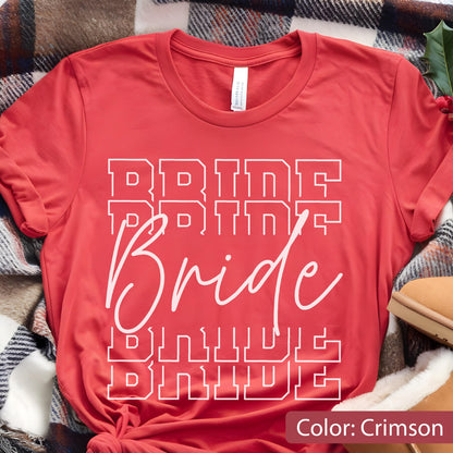 May include: Crimson red t-shirt with the word "Bride" in white, repeated vertically. The word "Bride" is in a cursive font, with the word "Bride" in a block font above and below. The shirt is folded on a plaid blanket.