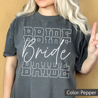 May include: Pepper-colored t-shirt with the word "Bride" in a white, bold, block letter font. The word "Bride" is repeated multiple times in a stacked design. The shirt has a relaxed fit and rolled-up sleeves.