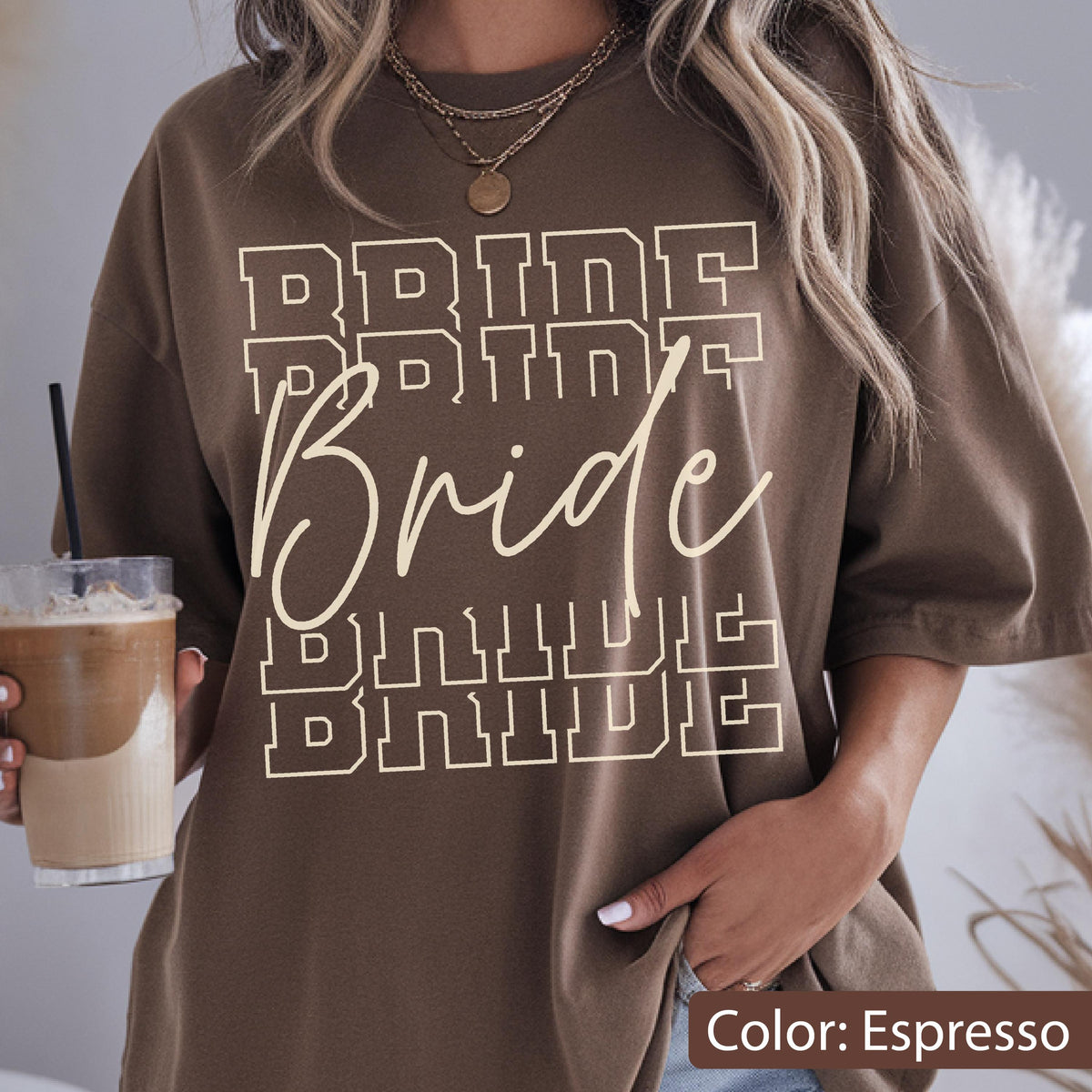 May include: Espresso-colored t-shirt with the word "Bride" in a layered design. The word "Bride" is in a cursive font, with the word "Bride" repeated in a block letter font above and below. The shirt is a casual style.