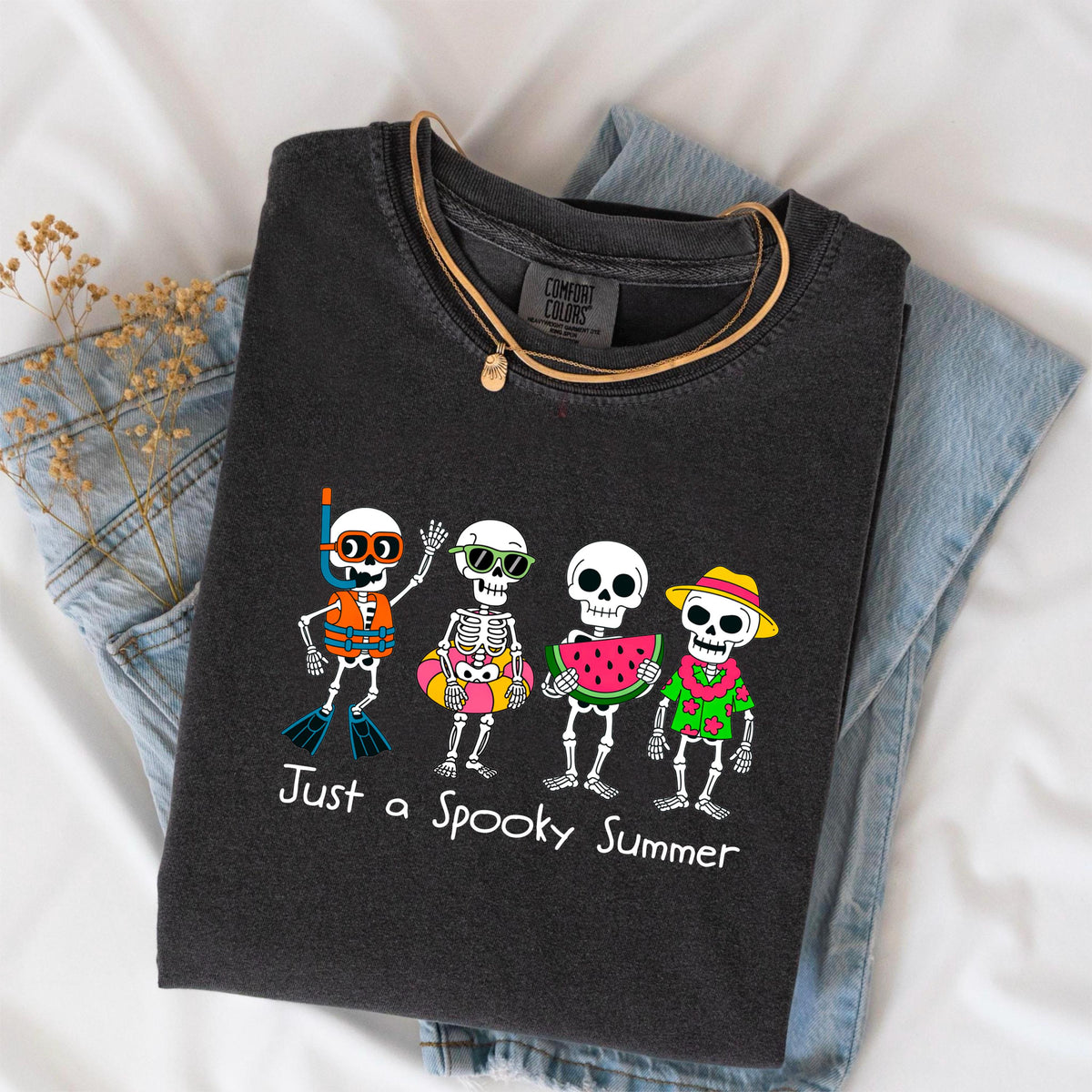 May include: Dark gray t-shirt with a graphic of four skeleton characters in summer attire. One skeleton wears a snorkel and life vest, another has sunglasses and a floatie, a third holds watermelon, and the last wears a Hawaiian shirt and hat. Text reads "Just a Spooky Summer."