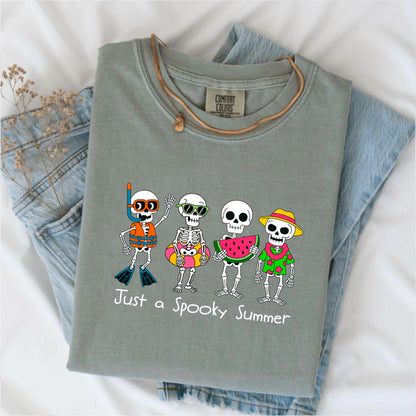 May include: Sage green Comfort Colors t-shirt featuring a graphic of four skeleton characters in summer attire. One wears a snorkel, another a floatie, a third holds watermelon, and the last wears a Hawaiian shirt. The text "Just a Spooky Summer" is below.