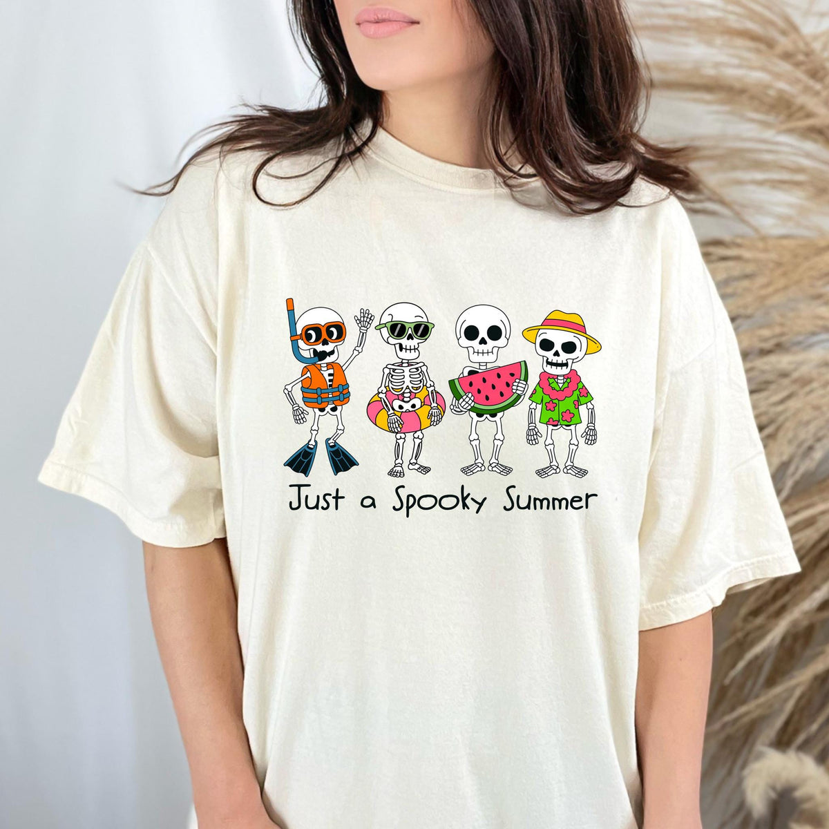 May include: Cream-colored t-shirt featuring a colorful graphic of four cartoon skeletons in summer attire. One skeleton wears a snorkel and life vest, another has sunglasses and a floatie, a third holds watermelon, and the last wears a hat and shirt. The text "Just a Spooky Summer" is below.