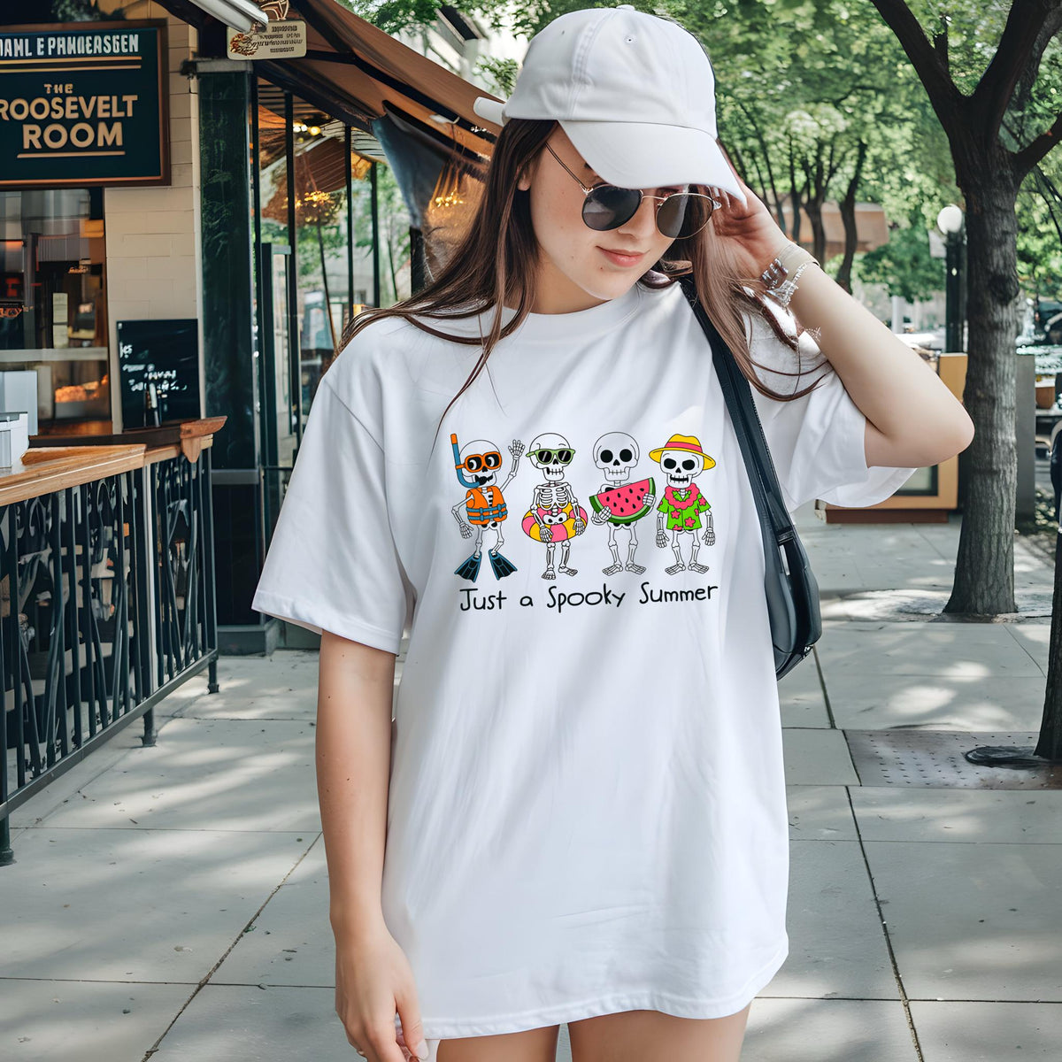 May include: White t-shirt featuring a colorful graphic of skeleton characters enjoying summer activities. The design includes skeletons with snorkeling gear, a life preserver, a watermelon slice, and a straw hat. The text "Just a Spooky Summer" is printed below the graphic.