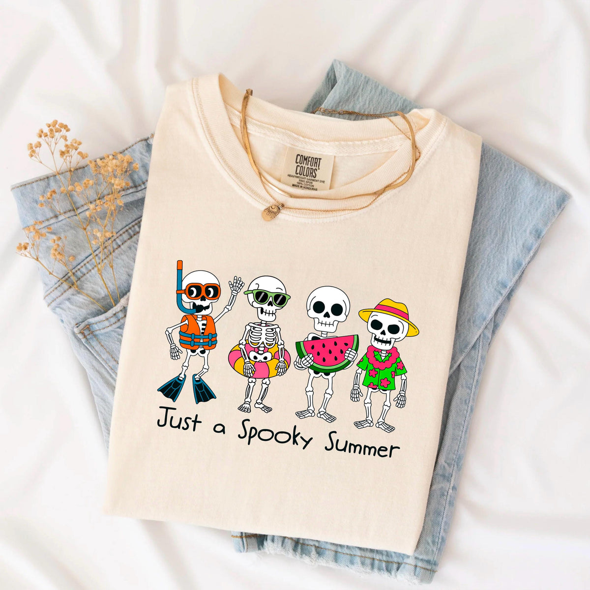 May include: Cream-colored t-shirt featuring a graphic of four skeleton characters in summer attire. One wears a snorkel and fins, another sunglasses and a floatie, a third holds watermelon, and the last wears a Hawaiian shirt and hat. The text "Just a Spooky Summer" is below.