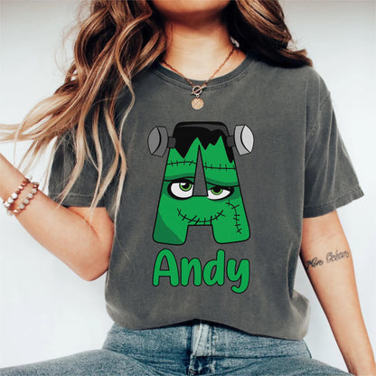 May include: A dark gray t-shirt featuring a green letter "A" designed as Frankenstein's monster, with bolts, stitches, and a black top. The name "Andy" is written in green below the letter. The shirt is a casual style.