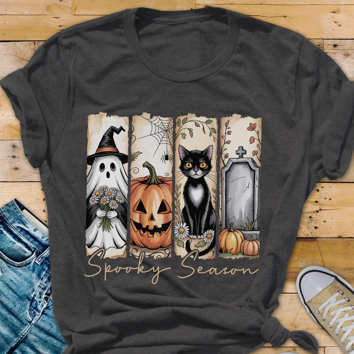 May include: A dark gray t-shirt featuring a graphic print divided into four vertical panels. The design displays a ghost in a witch hat, a jack-o'-lantern, a black cat, and a tombstone with pumpkins. The text Spooky Season is printed below.