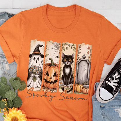 May include: An orange short-sleeve t-shirt featuring a graphic print of four vertical panels. The panels display a ghost in a witch hat holding flowers, a jack-o'-lantern, a black cat, and a tombstone. The text Spooky Season is printed below.