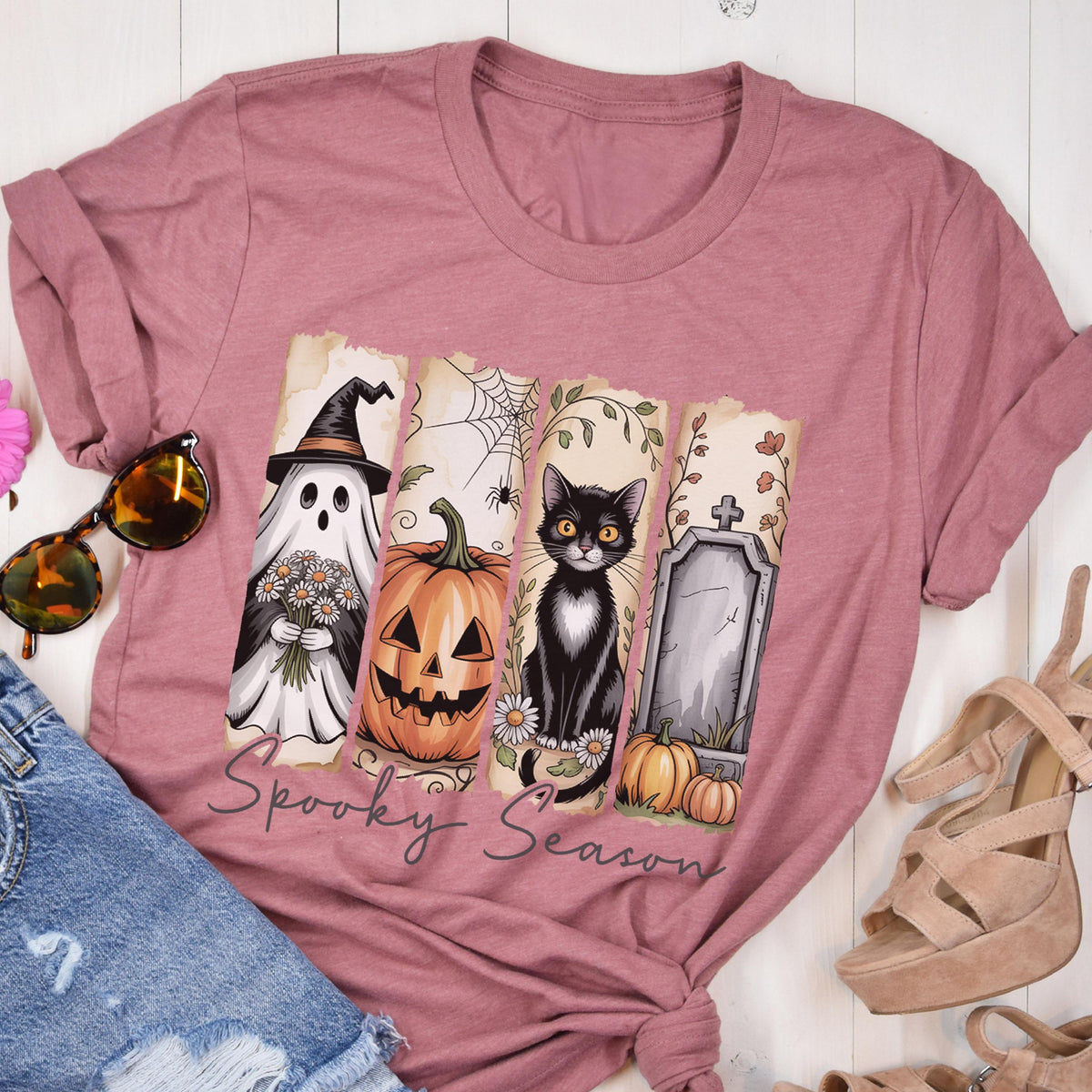 May include: A mauve short-sleeve t-shirt featuring a graphic print of four vertical panels with a ghost holding flowers, a jack-o'-lantern, a black cat, and a tombstone with pumpkins. The text Spooky Season is printed in black cursive below.