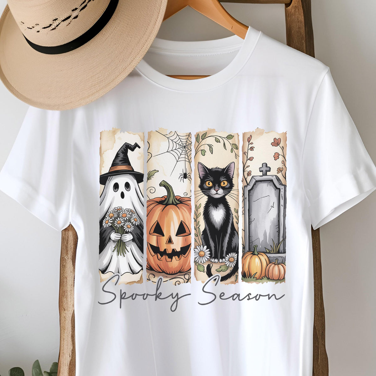 May include: A white short-sleeve t-shirt featuring a graphic print with four vertical panels depicting a ghost with flowers, a jack-o'-lantern, a black cat, and a tombstone with pumpkins. The text Spooky Season is printed below in gray.