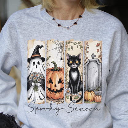 May include: A light gray crewneck sweatshirt featuring a graphic print of four vertical panels with a ghost holding flowers, a jack-o'-lantern, a black cat, and a tombstone. The text Spooky Season is printed in a cursive font below the panels.