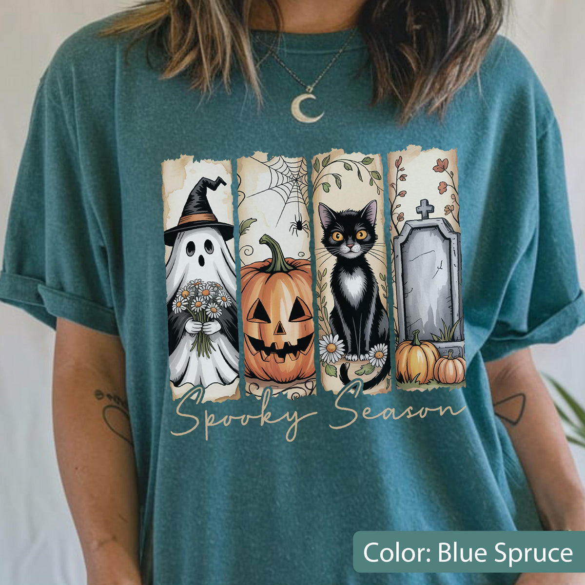 May include: Blue spruce t-shirt with a Halloween-themed graphic. The design features a ghost, jack-o'-lantern, black cat, and tombstone. The words "Spooky Season" are written in a script font across the bottom of the graphic.