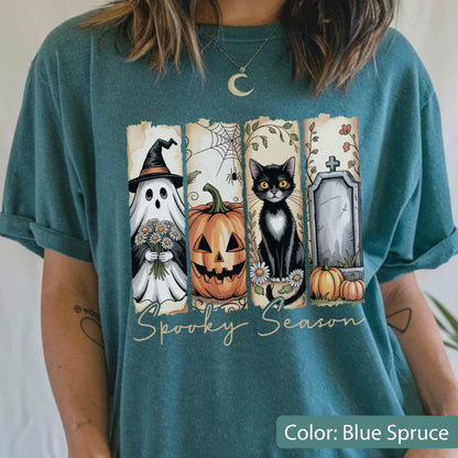 May include: Blue spruce t-shirt with a Halloween-themed graphic. The design features a ghost, jack-o'-lantern, black cat, and tombstone. The words "Spooky Season" are written in a script font across the bottom of the graphic.