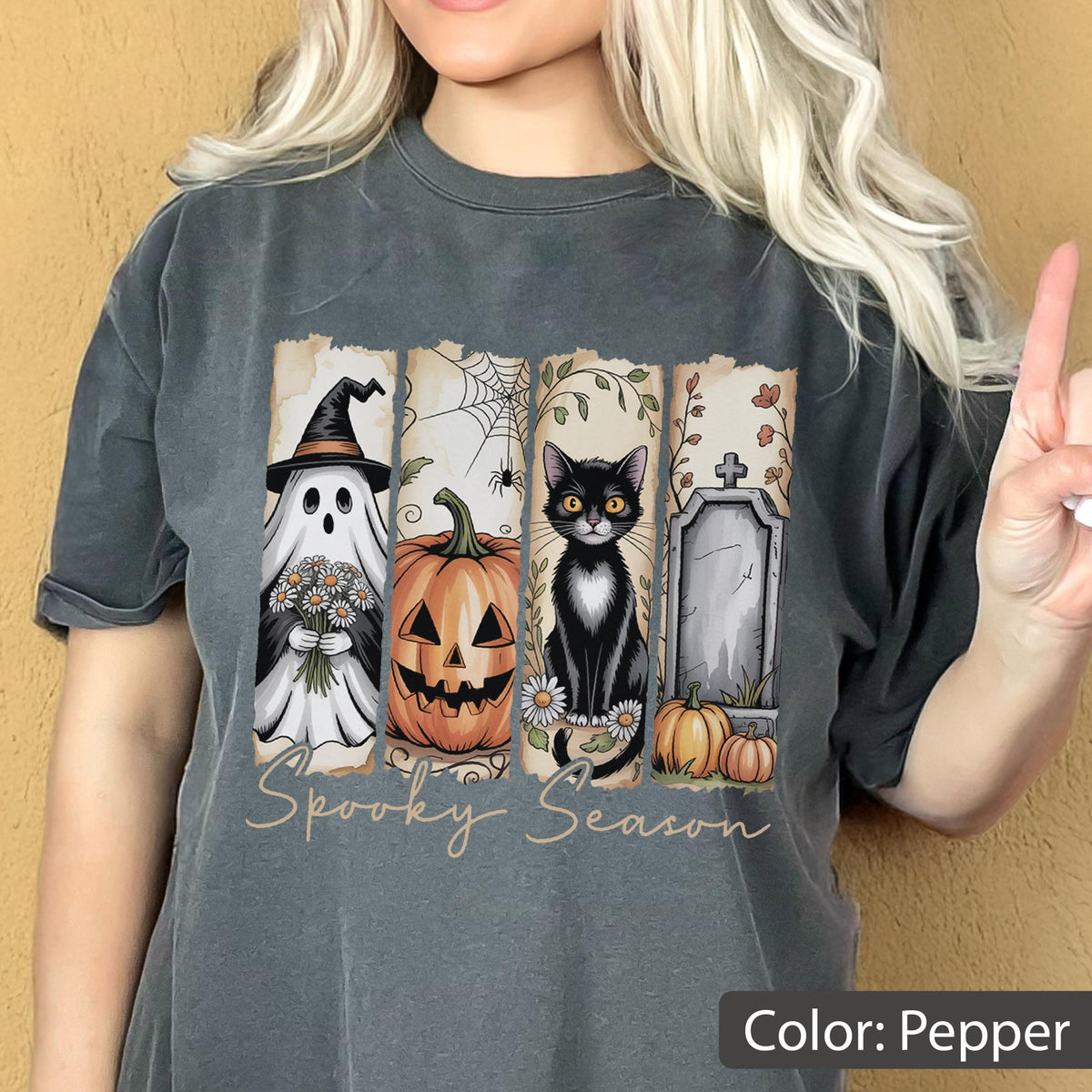 May include: Pepper-colored t-shirt with a Halloween-themed graphic. The design features a ghost, jack-o'-lantern, black cat, and tombstone. The words "Spooky Season" are written in a cursive font across the bottom of the graphic.