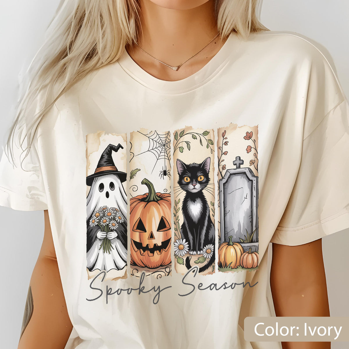 May include: Ivory-colored t-shirt featuring a Halloween-themed design. The graphic includes a ghost in a witch hat, a carved pumpkin, a black cat, and a tombstone. The words "Spooky Season" are written in a script font below the images.
