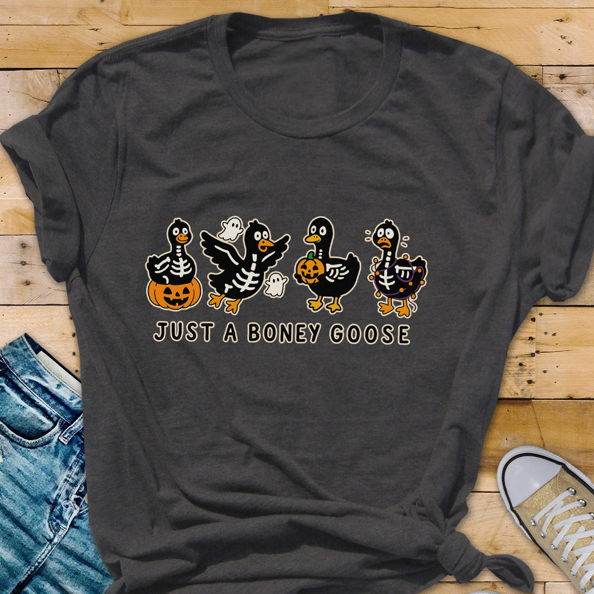May include: Dark gray t-shirt featuring a Halloween-themed graphic. The design includes four cartoon geese dressed as skeletons, holding pumpkins and accompanied by ghosts. The text "JUST A BONEY GOOSE" is printed below the geese.