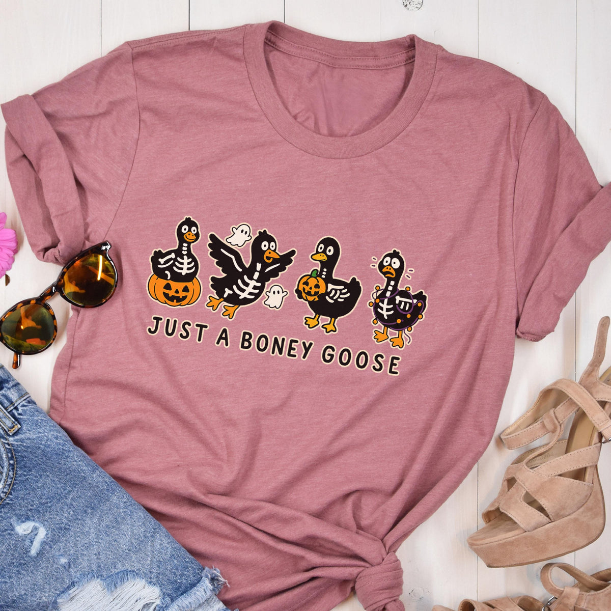 May include: Dusty rose t-shirt with a Halloween-themed graphic. The design features four cartoon geese with skeleton details, pumpkins, and ghosts. The text "JUST A BONEY GOOSE" is printed below the geese.