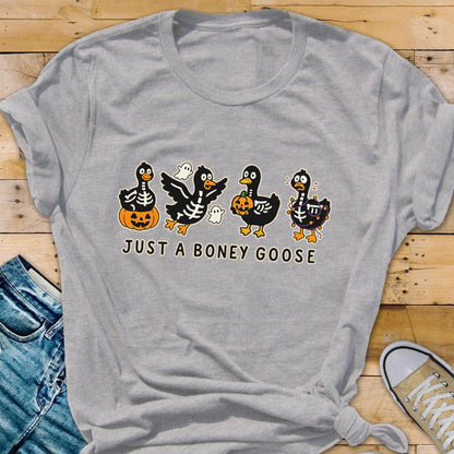 May include: A heather gray t-shirt featuring a Halloween-themed graphic. The design includes four cartoon geese dressed as skeletons, holding pumpkins and accompanied by ghosts. The text "JUST A BONEY GOOSE" is printed below the geese.