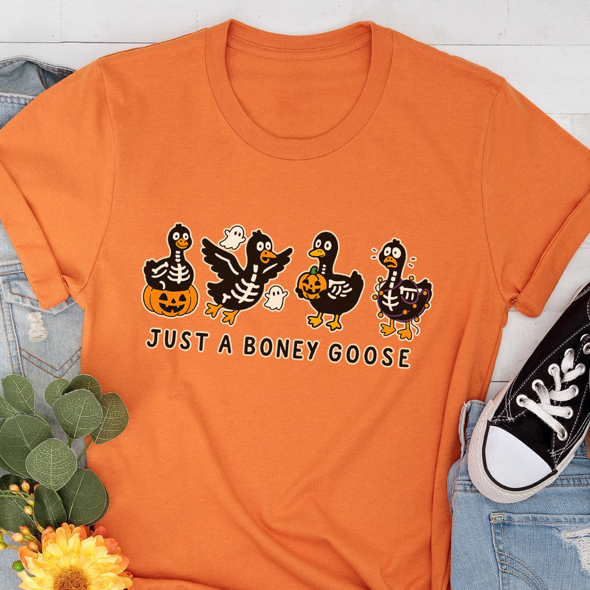 May include: Orange t-shirt featuring a Halloween-themed graphic of four cartoon geese in skeleton costumes, with pumpkins and ghosts. The text "JUST A BONEY GOOSE" is printed below the design. The shirt is paired with blue jeans and a black sneaker.