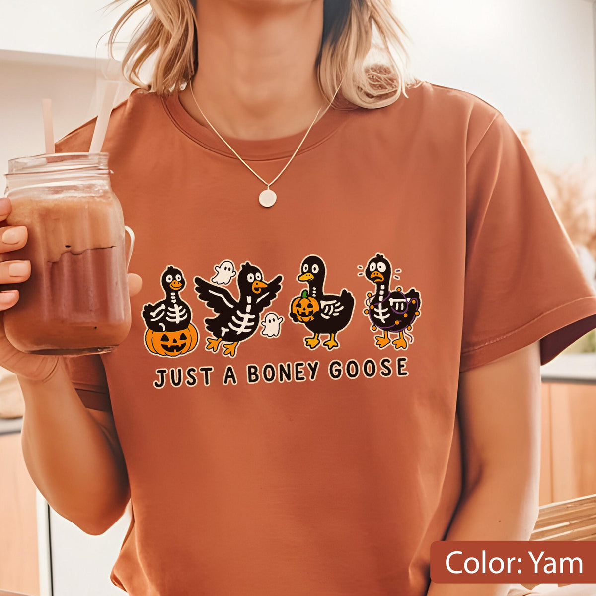 May include: A yam-colored t-shirt featuring a Halloween-themed graphic. The design includes four cartoon geese with skeleton-like bodies, pumpkins, and ghosts. The text "JUST A BONEY GOOSE" is printed below the geese.