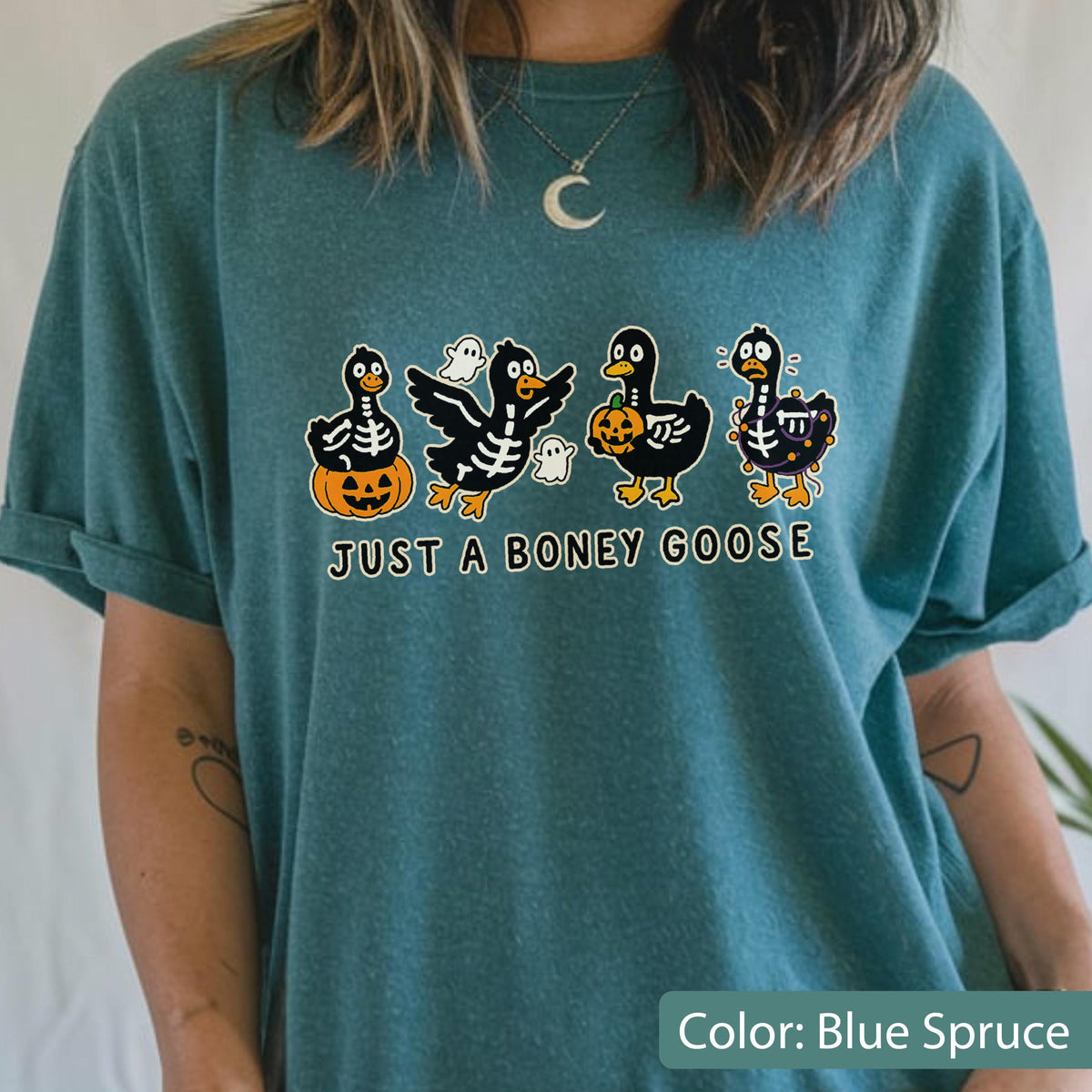 May include: Blue spruce t-shirt with a Halloween-themed graphic. The design features four cartoon geese in skeleton costumes, holding pumpkins and accompanied by ghosts. Text reads "JUST A BONEY GOOSE".