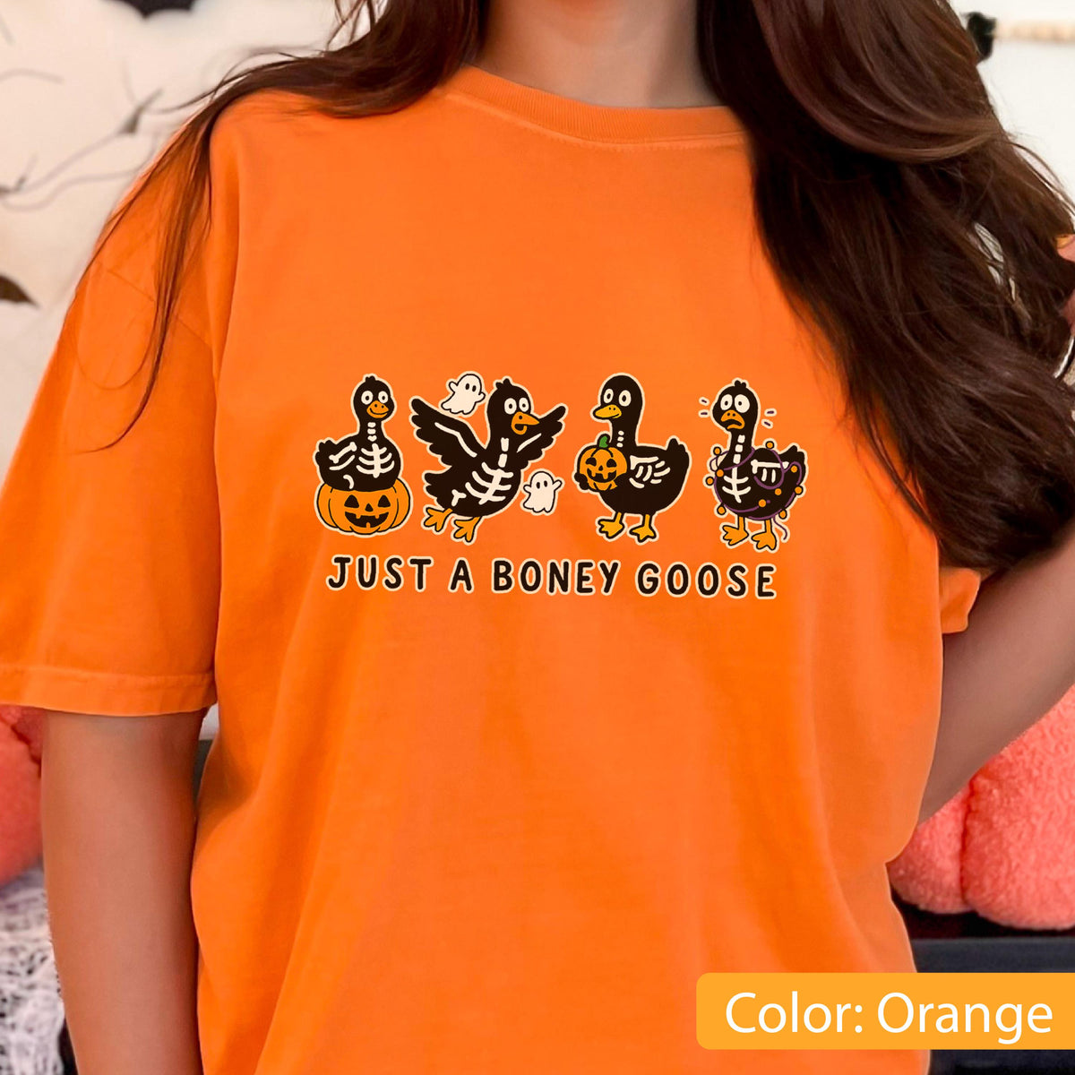 May include: Orange t-shirt featuring a Halloween-themed graphic. The design includes four cartoon geese with skeleton and pumpkin details, along with the text "JUST A BONEY GOOSE". The shirt is a solid orange color.