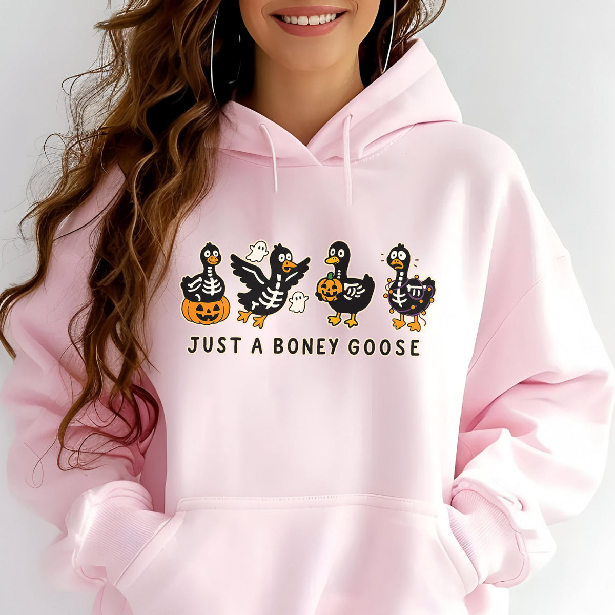 May include: A light pink hoodie with a graphic of four cartoon geese dressed as skeletons, holding pumpkins and a ghost. The text "JUST A BONEY GOOSE" is printed below the geese in white.