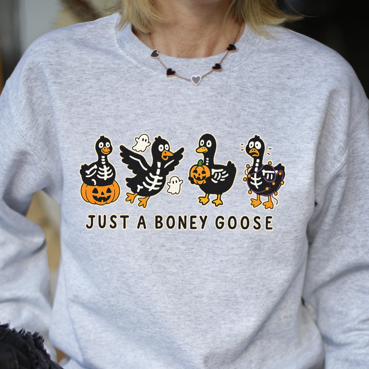 May include: A light gray sweatshirt featuring a Halloween-themed graphic. The design includes four cartoon ducks with skeleton details, pumpkins, and ghosts. The text "JUST A BONEY GOOSE" is printed below the ducks.