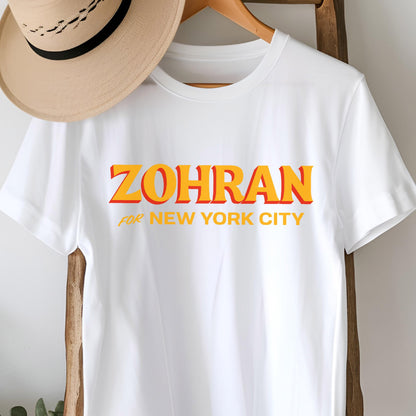 May include: A white short-sleeved t-shirt featuring the text ZOHRAN in large orange and yellow letters with a red drop shadow, followed by the text FOR NEW YORK CITY in yellow. A tan fedora hat with a black band is positioned above the t-shirt.