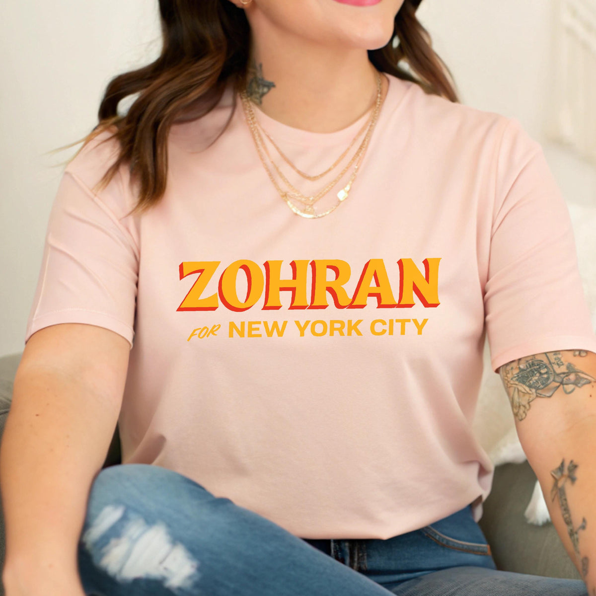 May include: A light pink short-sleeved t-shirt featuring the text ZOHRAN in large orange letters with a yellow outline, followed by the text FOR NEW YORK CITY in smaller yellow letters centered on the chest of the garment.
