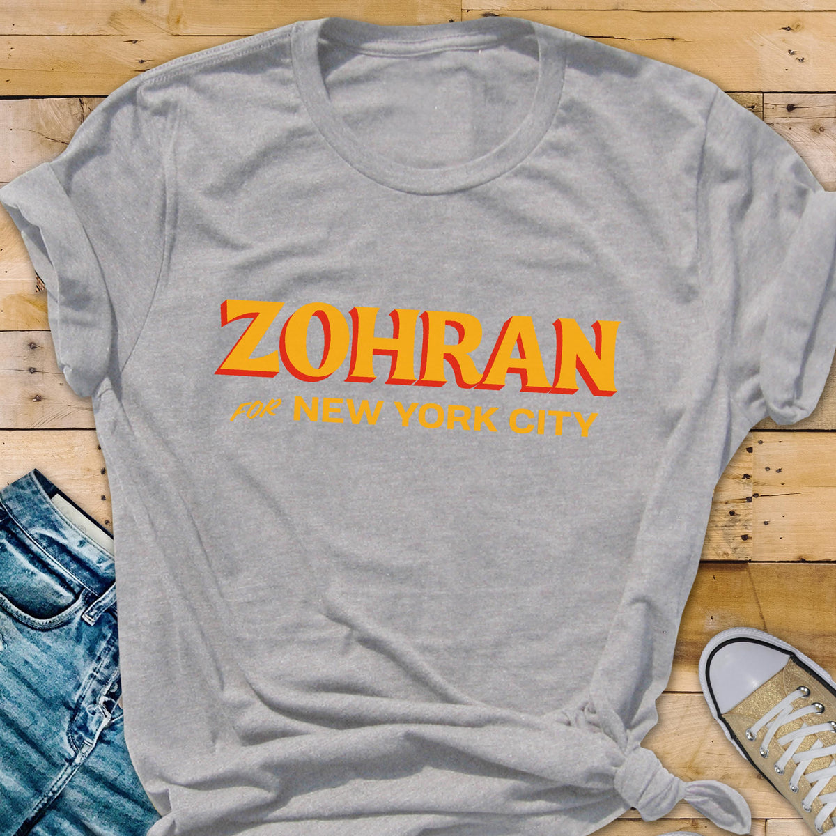 May include: A light gray crew neck t-shirt featuring the text ZOHRAN in bold orange letters with a red shadow effect, followed by the text for NEW YORK CITY in smaller yellow letters centered on the front of the garment.