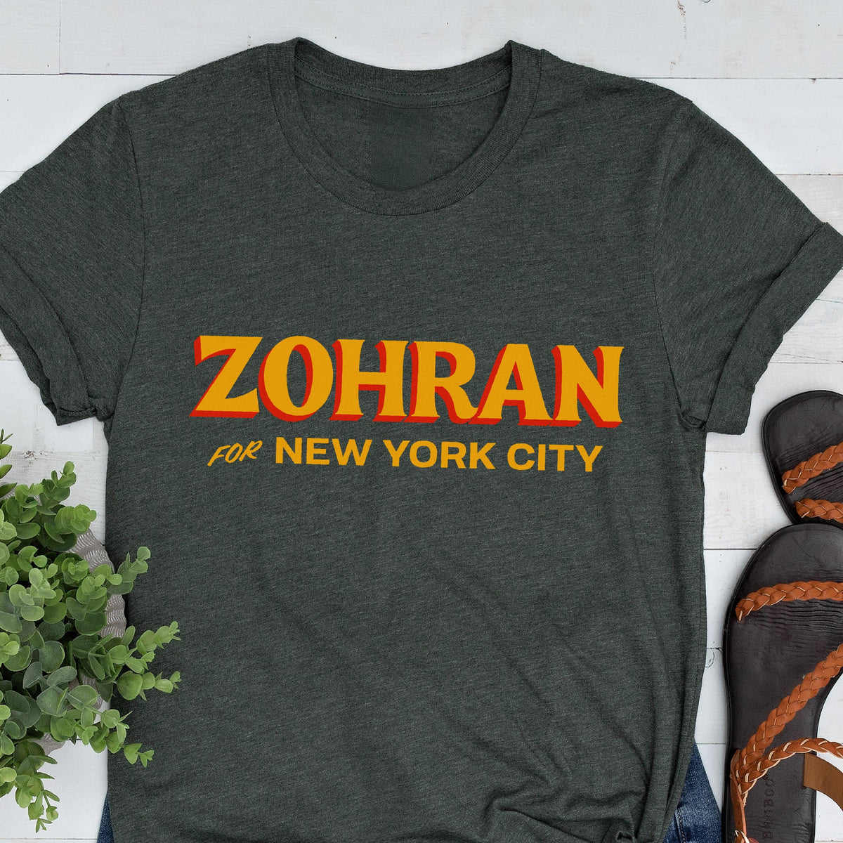 May include: A dark charcoal gray short-sleeved t-shirt featuring the text ZOHRAN in large yellow letters with a red outline, followed by the text FOR NEW YORK CITY in smaller yellow letters centered on the front of the garment.