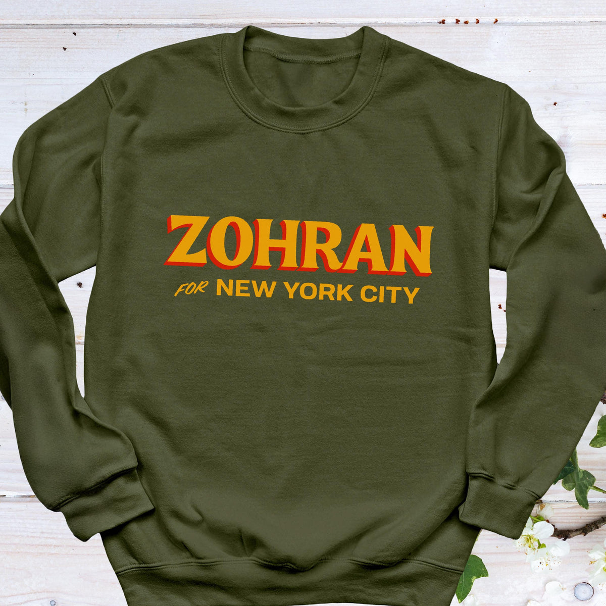 May include: Olive green sweatshirt with the name "ZOHRAN" in orange block letters with a red outline. Below the name, the text "FOR NEW YORK CITY" is printed in orange. The sweatshirt is a crew neck style.
