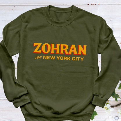 May include: Olive green sweatshirt with the name "ZOHRAN" in orange block letters with a red outline. Below the name, the text "FOR NEW YORK CITY" is printed in orange. The sweatshirt is a crew neck style.