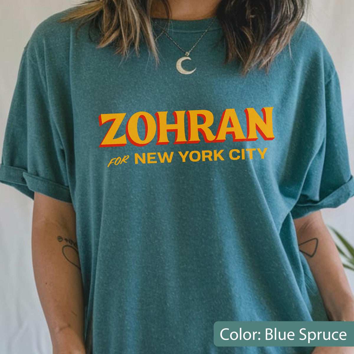 May include: Blue spruce t-shirt with the word "ZOHRAN" in orange letters with a red outline, and the words "FOR NEW YORK CITY" in smaller, yellow letters. The shirt has rolled-up sleeves.