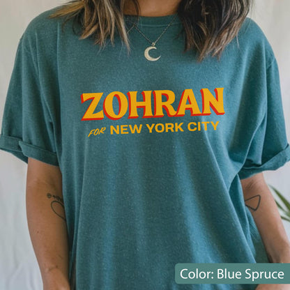 May include: Blue spruce t-shirt with the word "ZOHRAN" in orange letters with a red outline, and the words "FOR NEW YORK CITY" in smaller, yellow letters. The shirt has rolled-up sleeves.