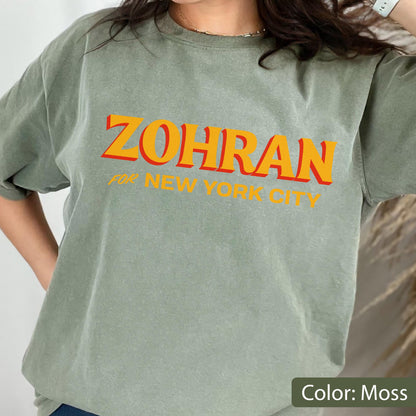 May include: A moss green t-shirt with the word "ZOHRAN" in orange letters with a red outline, and the text "FOR NEW YORK CITY" in smaller orange letters. The shirt is a solid color and has short sleeves.