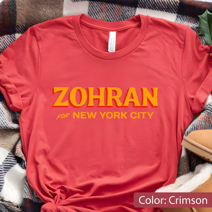 May include: Crimson-colored t-shirt with the word "ZOHRAN" in large, orange letters, and "FOR NEW YORK CITY" in smaller orange letters. The shirt is made of a soft material and is displayed on a plaid blanket.
