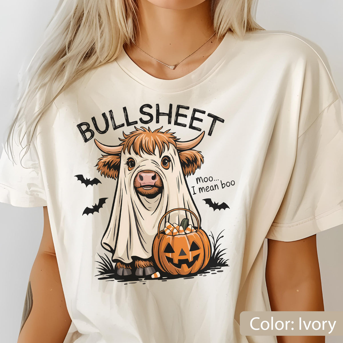 May include: An ivory t-shirt featuring a graphic of a highland cow dressed as a ghost, holding a jack-o'-lantern bucket filled with candy corn. The text reads BULLSHEET and moo... I mean boo, accompanied by black bat silhouettes.