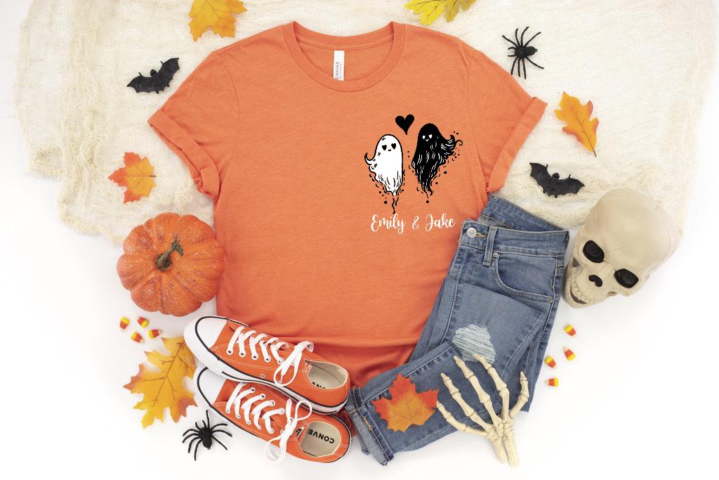 May include: An orange short-sleeved t-shirt featuring a graphic of two ghosts, one white and one black, with a small black heart between them. Below the ghosts, the text reads Emily & Jake in a white script font. The shirt has a crew neckline.