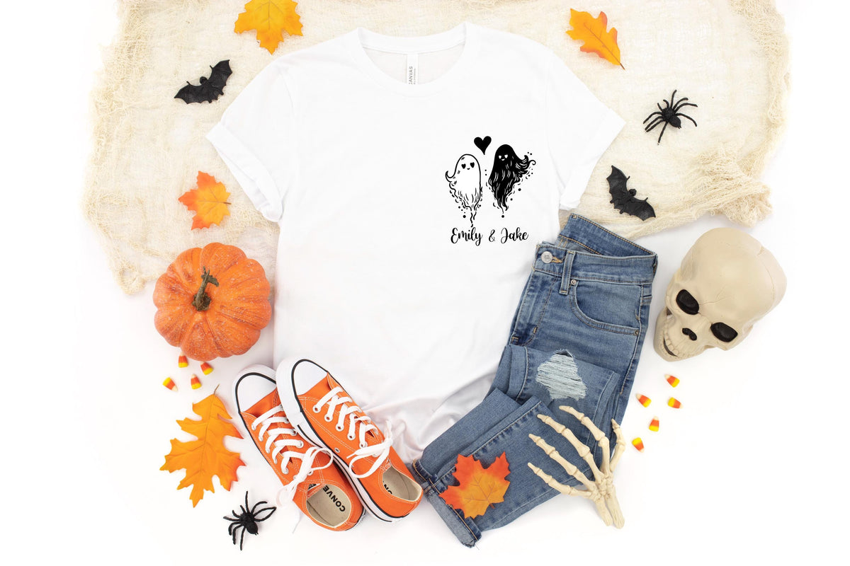May include: A white short-sleeve t-shirt featuring a black graphic of two ghosts with a heart and the text Emily & Jake. The shirt is paired with blue denim jeans, orange canvas sneakers, a pumpkin, a skull, and decorative autumn leaves.