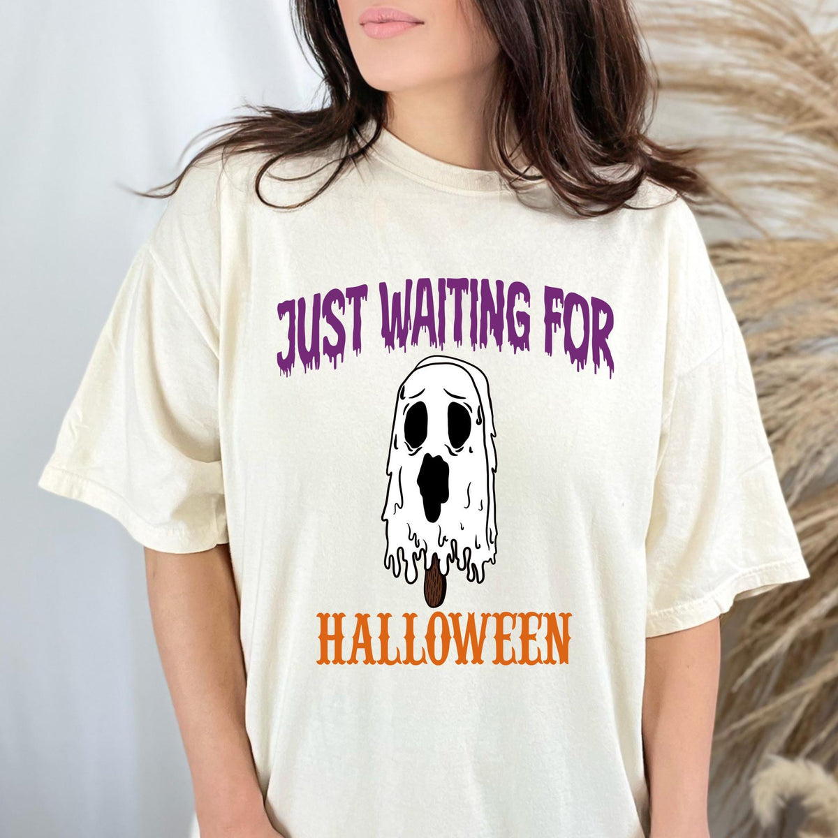 May include: Cream-colored t-shirt with the text "JUST WAITING FOR HALLOWEEN." The shirt features a graphic of a melting ghost popsicle. The text is in purple and orange. The ghost has a sad expression.