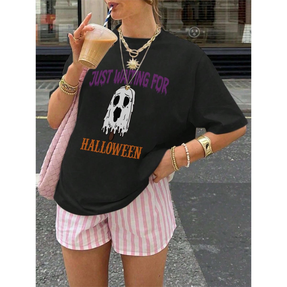 May include: Black t-shirt with the text "JUST WAITING FOR HALLOWEEN" and a ghost graphic. The shirt is paired with pink and white striped shorts. The person is wearing gold jewelry and holding a drink.