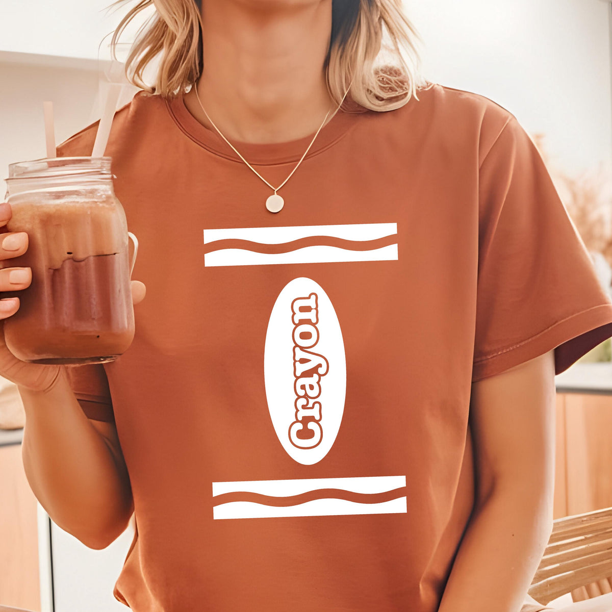 May include: A rust-colored short-sleeve t-shirt featuring a white graphic design on the front. The design consists of two horizontal wavy lines and a central vertical oval containing the word Crayon in a bold, white, sans-serif typeface.