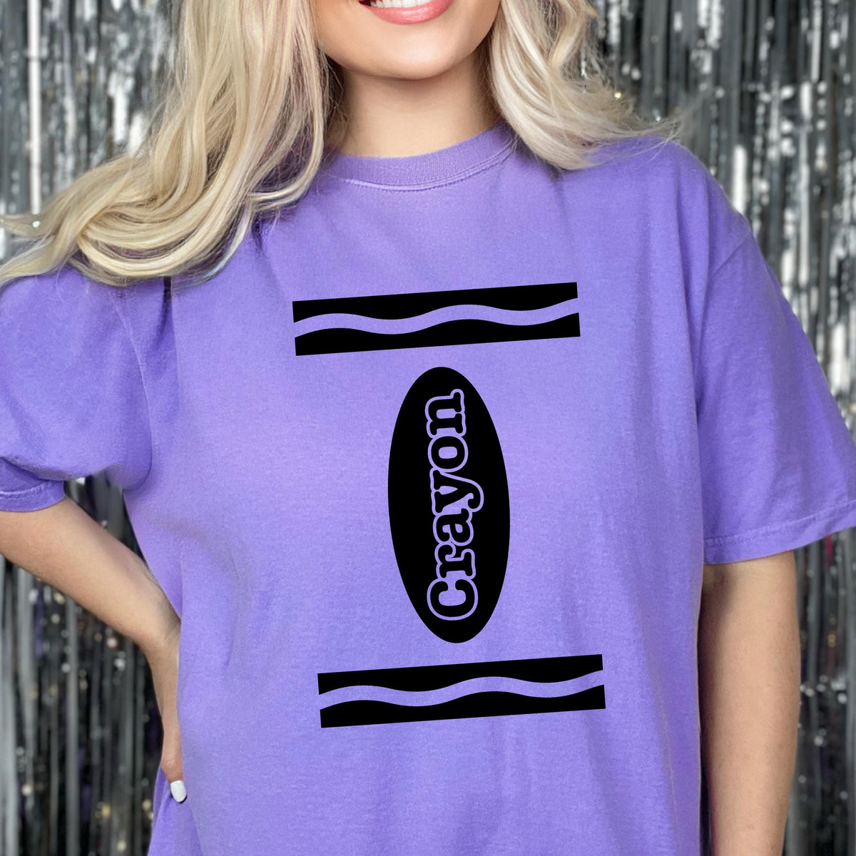 May include: A light purple short-sleeve t-shirt featuring a black graphic design that mimics a crayon wrapper. The design includes two horizontal wavy black lines and a central black oval containing the word Crayon in light purple text.