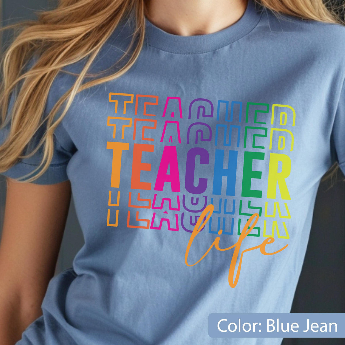 May include: A blue jean colored t-shirt featuring a multi-colored graphic print. The design displays the word TEACHER repeated in stacked, colorful block letters, with the word life written in a cursive orange font underneath the text.