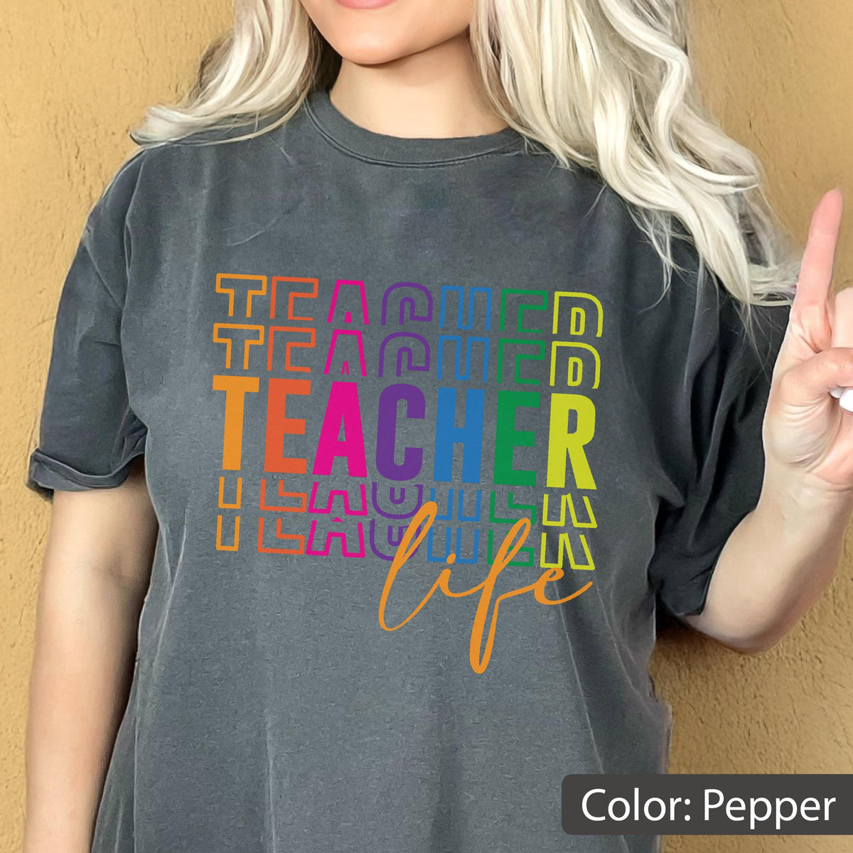 May include: A pepper-colored t-shirt featuring the word TEACHER repeated in a stacked, multi-colored block font with the word life written in a cursive orange script underneath. The design uses vibrant shades of orange, pink, purple, blue, and yellow.