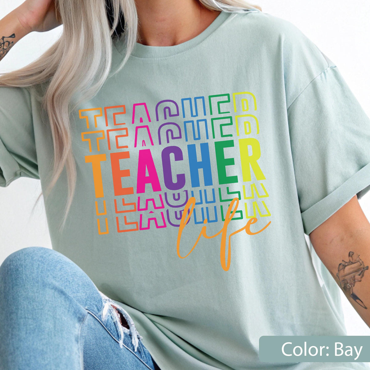 May include: A light sage green short-sleeve t-shirt featuring a multi-colored graphic print. The design displays the word TEACHER in bold, stacked, colorful letters, with the word life written in a cursive orange script underneath the text.